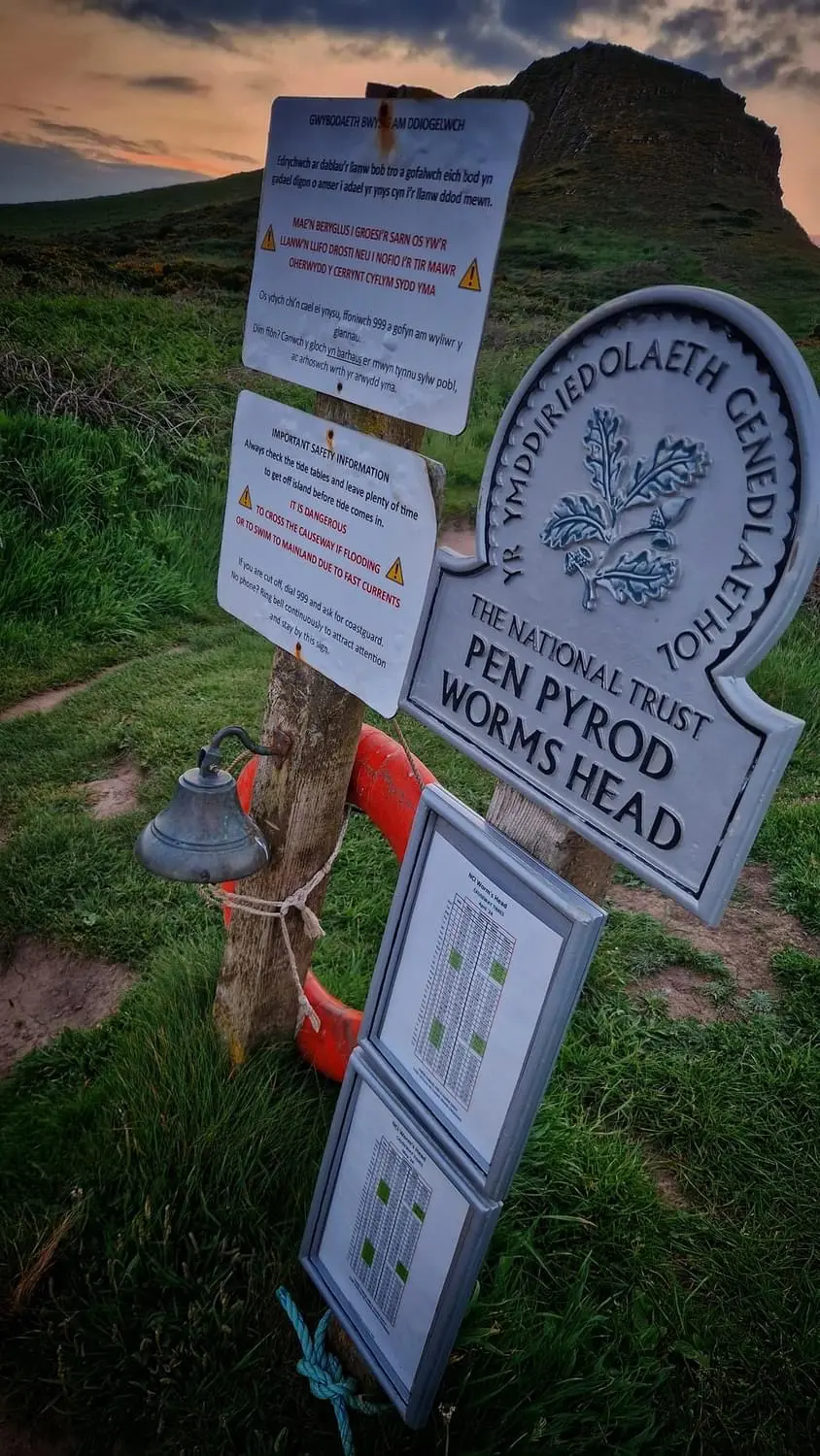 The emergency bell, life ring, safety signs and causeway tide tables at the Worms Head landing point, with the Inner Head behind at sunset
