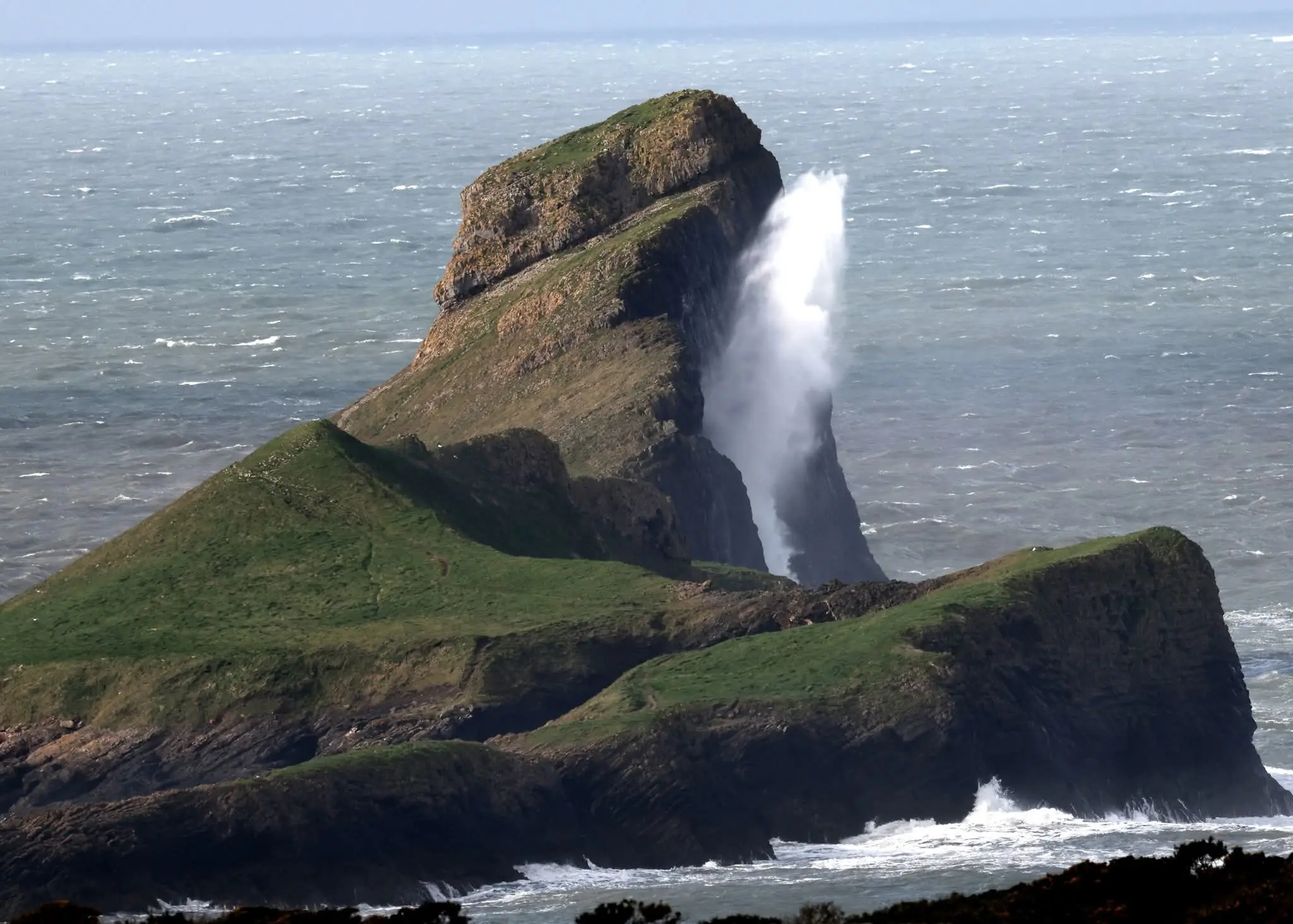 A tall plume of white sea spray shooting up from the blowhole on the Outer Head of Worms Head