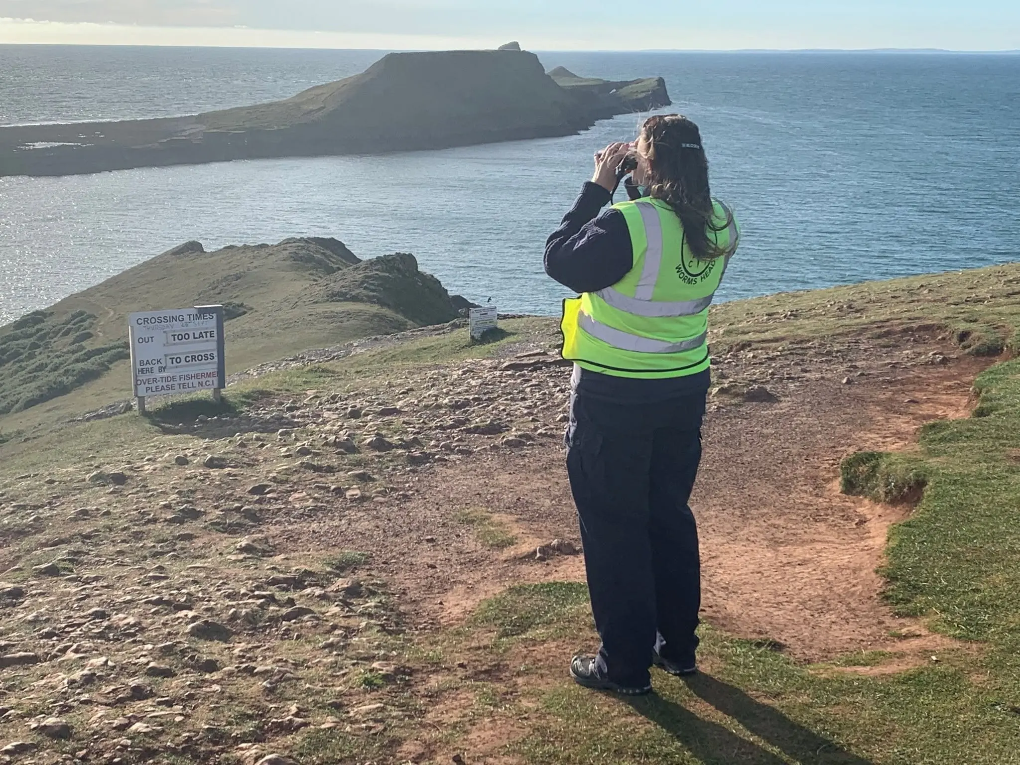 A watchkeeper using binoculars beside the causeway crossing times board, with Worms Head behind