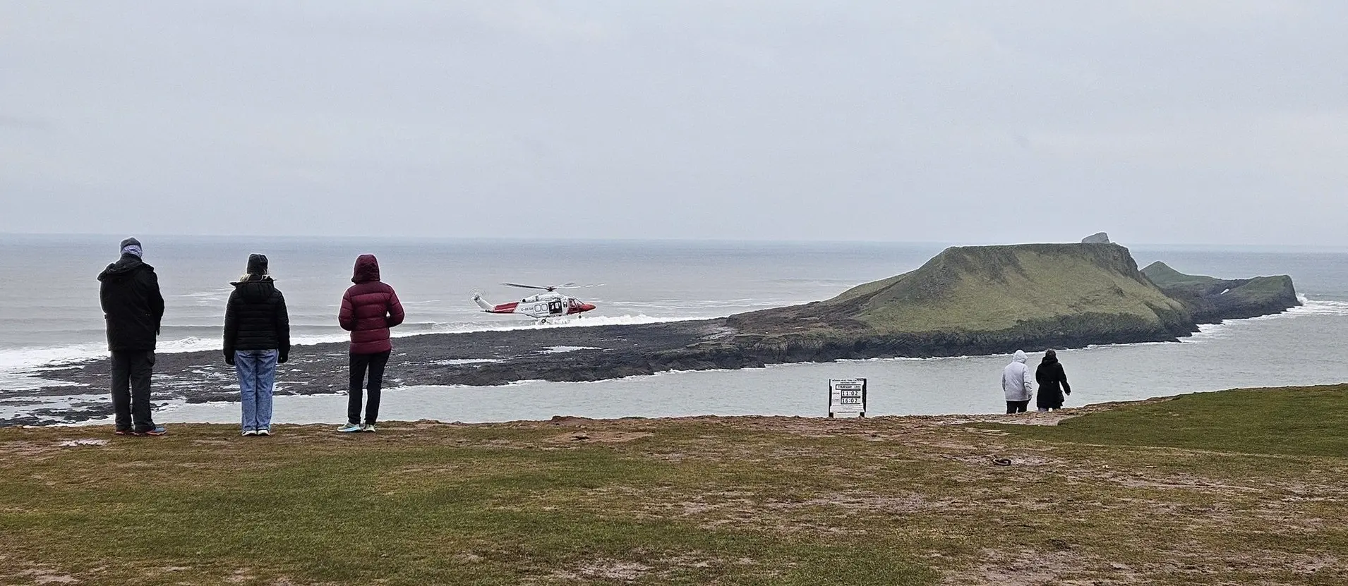 Onlookers on the clifftop watching a Coastguard helicopter operating near the Worms Head causeway during a real rescue