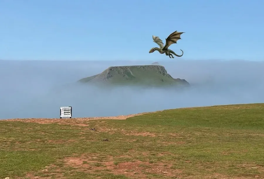 A green dragon flying over Worms Head as the island rises out of a bank of sea mist, seen from the clifftop