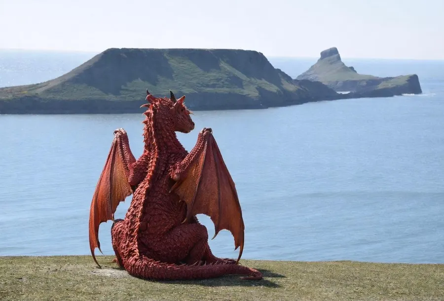 A big red Welsh dragon sitting on the clifftop with its wings folded, gazing out at Worms Head across the sea