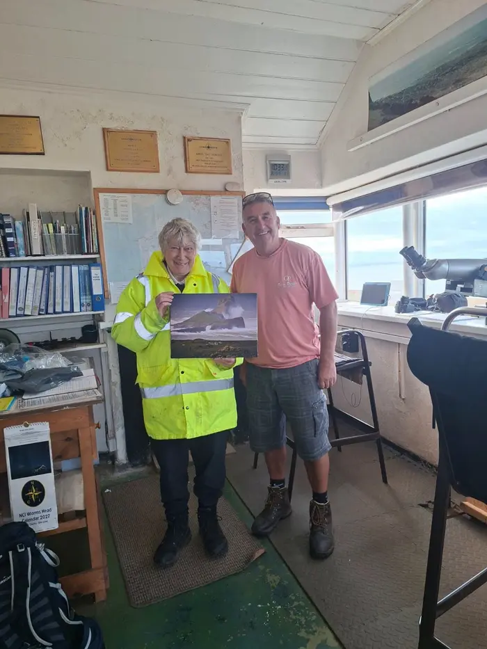 Photographer Gary Williams in the lookout's watch room with a watchkeeper, holding his canvas print of Worms Head with waves breaking below it