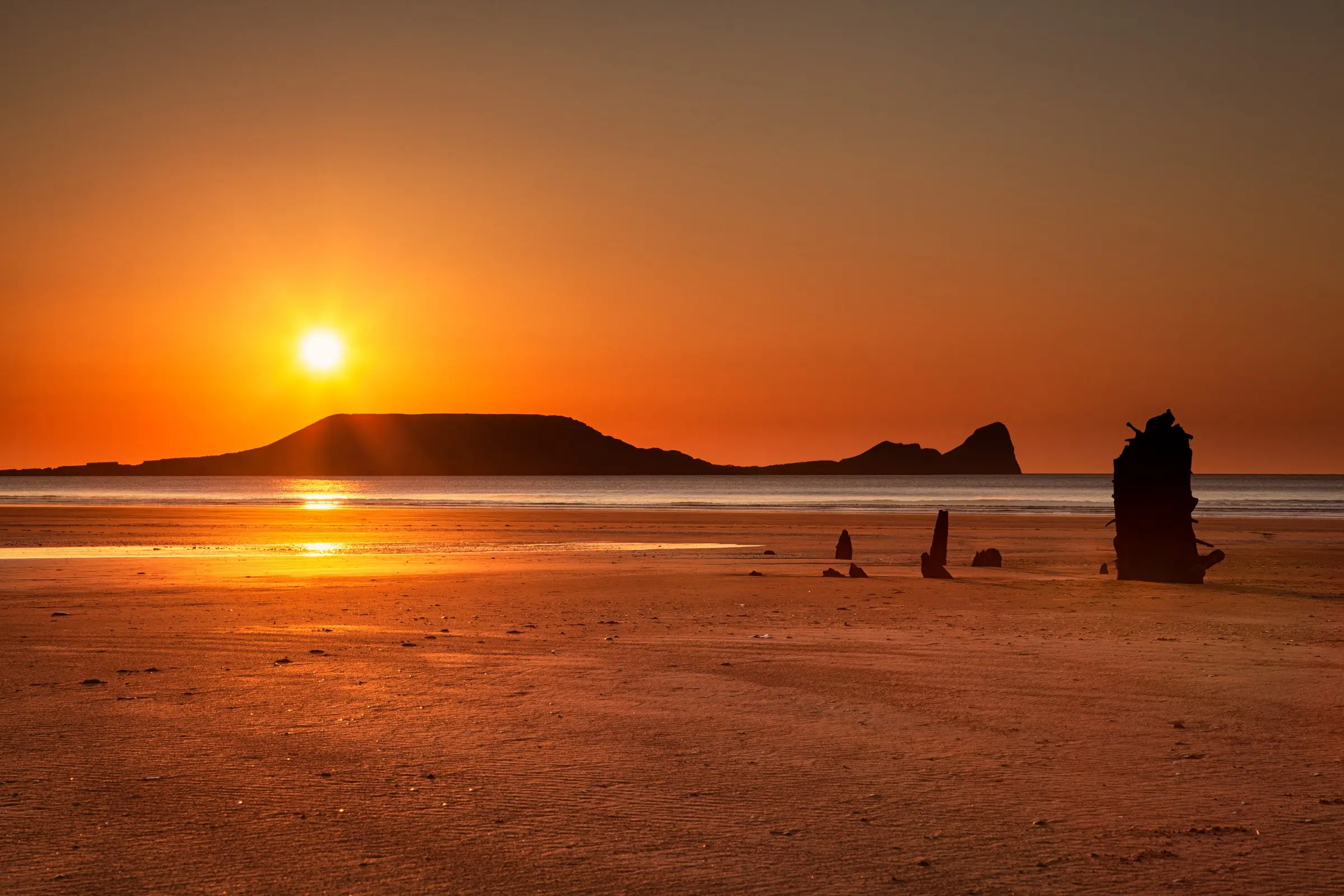The ribs of the Helvetia wreck on Rhosili beach at sunset, with the sun setting beside Worms Head
