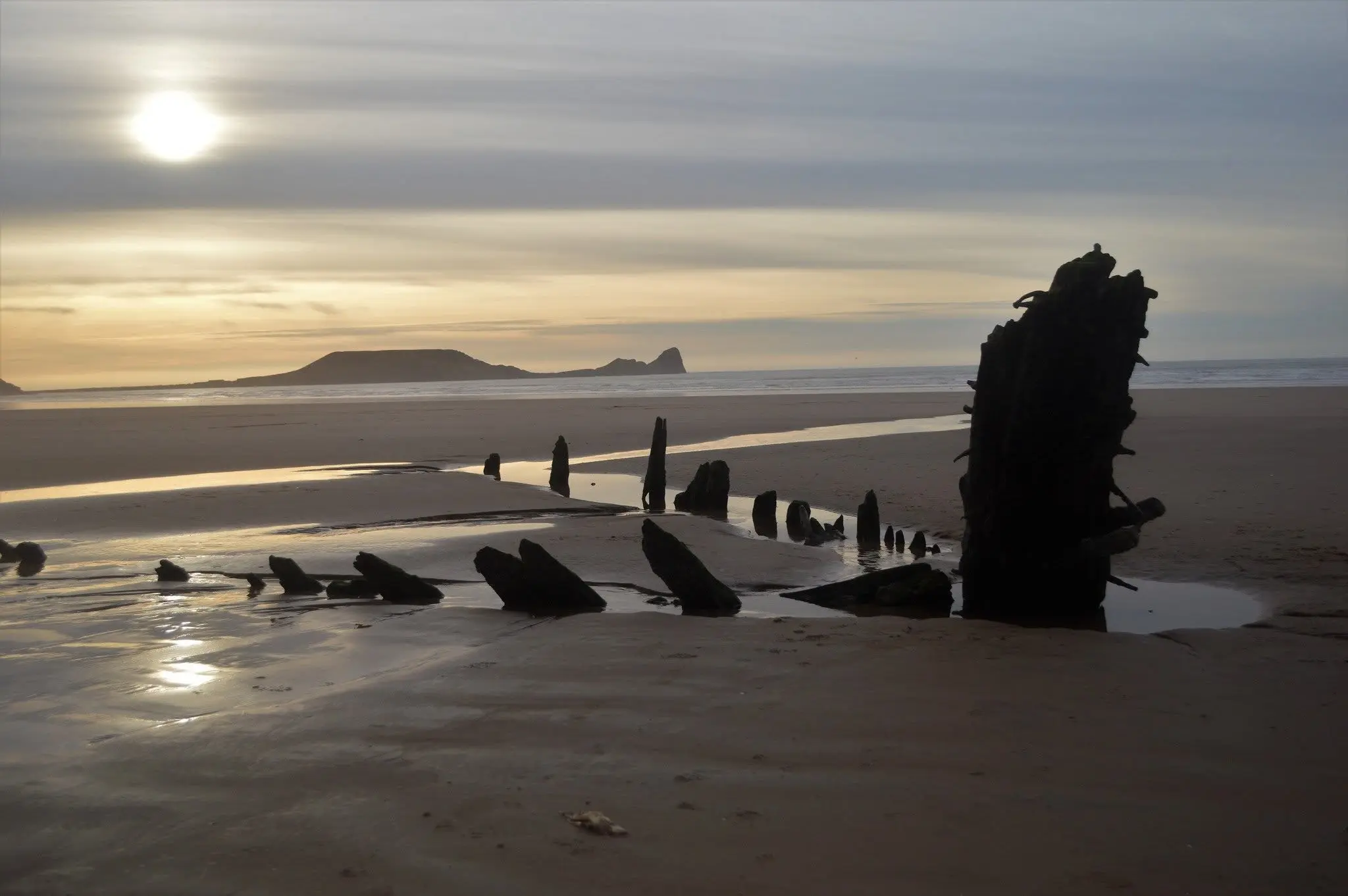 The historic oak timbers of the Helvetia shipwreck on Rhosili beach at sunset, with Worms Head in the distance