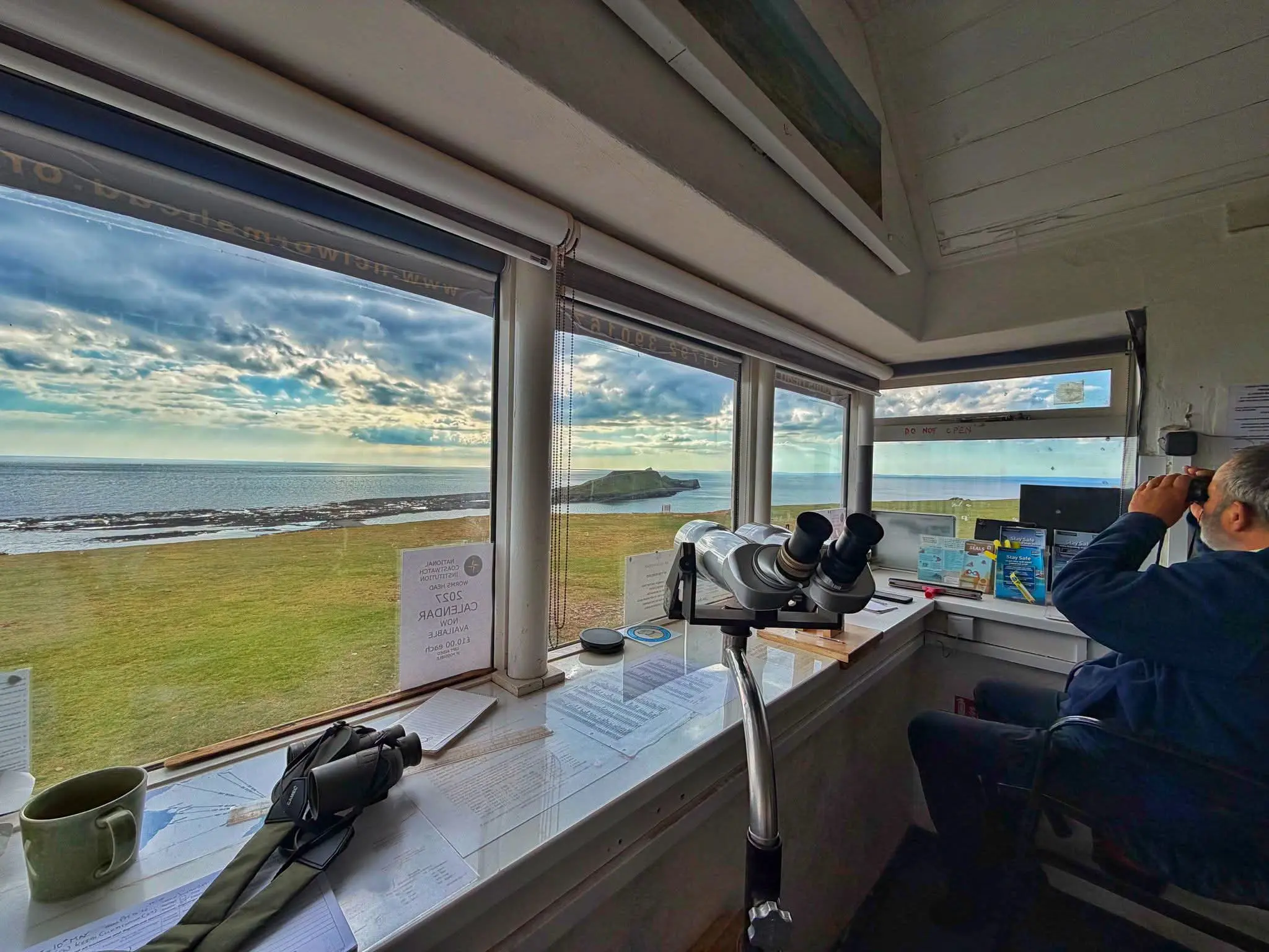 Inside the NCI Worms Head lookout: a watchkeeper scans the sea through binoculars, with the causeway and Worms Head through the window