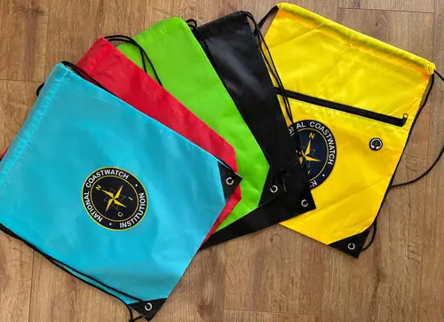 Five NCI-branded drawstring cinch bags in teal, red, green, black, and yellow, each printed with the National Coastwatch Institution compass badge