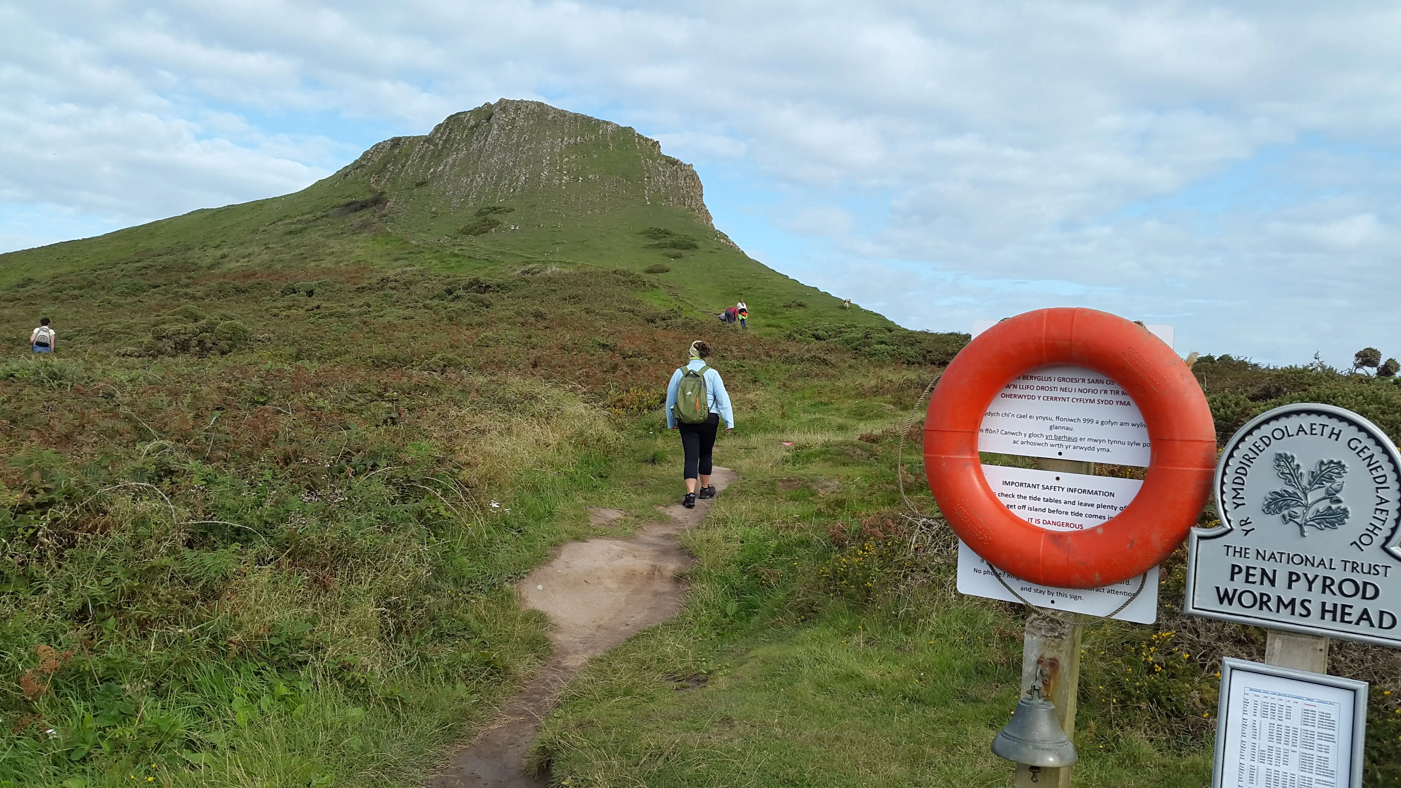 A walker arriving at the landing point onto Worms Head, with the causeway safety sign, emergency bell, and life ring alongside the path