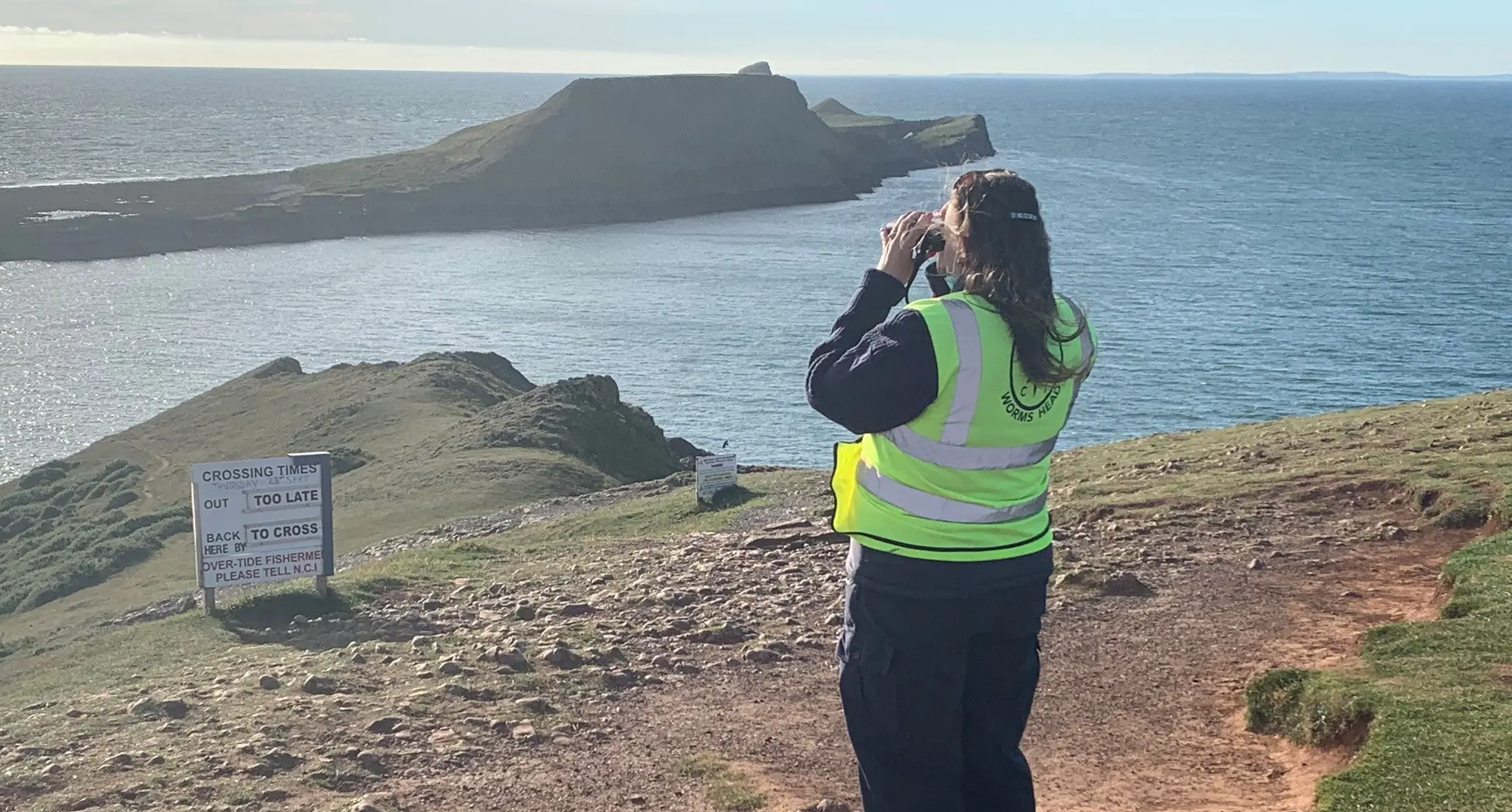 An NCI Worms Head watchkeeper scanning the causeway through binoculars beside the crossing-times board, with Worms Head beyond