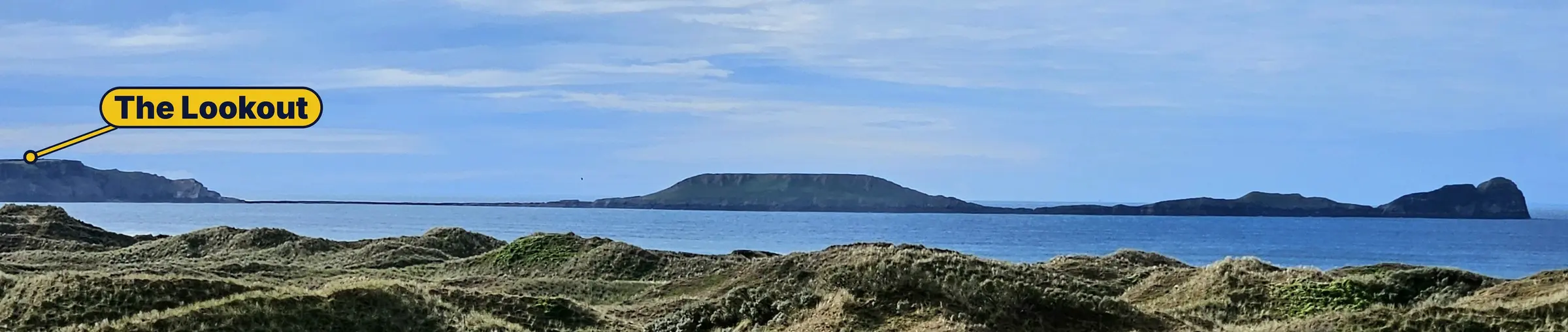 Worms Head stretching out to sea, seen across the grassy sand dunes of Rhosili Bay, with the NCI lookout marked on the headland to the left